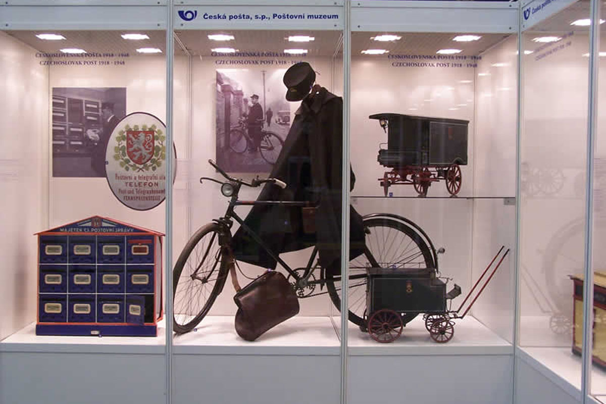 Vitrína v Poštovním muzeu s cyklistickým kolem, uniforma poštovního doručovatele, makety poštovních vozů a poštovní schránka