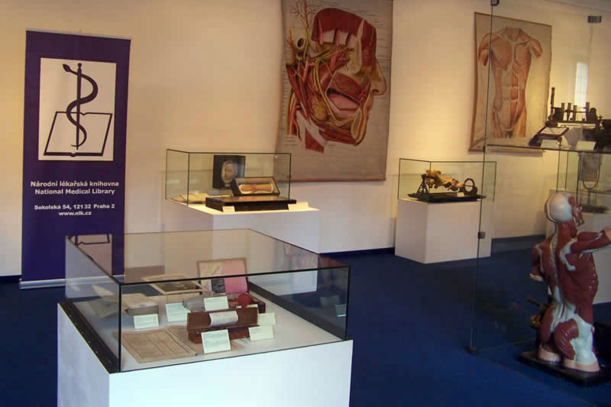 Národní lékařská knihovna s naaranžovanými exponáty, vitríny, roletka s logem knihovny a obrázky na stěnách